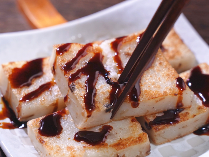 Several white radish cakes served on a plate. They have been fried golden brown with sweet soy sauce drizzled across them. 