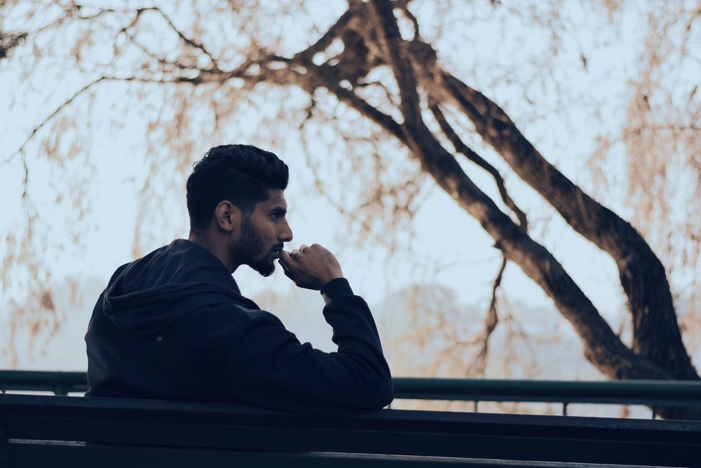 A side profile of man sitting on a park bench. His hand is to his mouth and his facial expression is that of contemplation. 