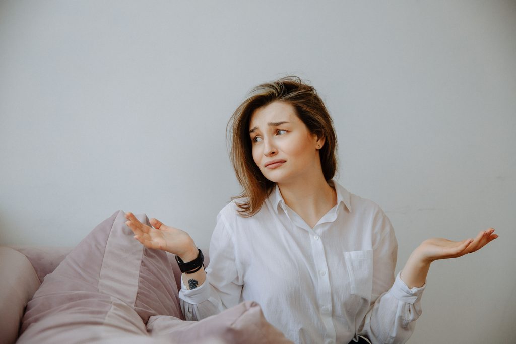 Woman in white shirt sitting indoors on a couch, gesturing with a confused expression, hands up in a shrug.